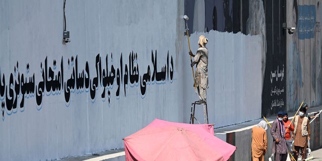A man paints over murals on a concrete wall with a message reading "For an Islamic system and independence, you have to go through tests and stay patient", in Kabul.