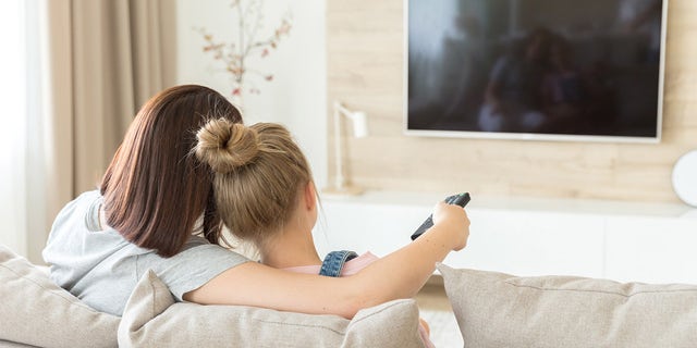 A mother and daughter are shown sitting on a sofa watching TV. Situations such as the invasion of Ukraine can be magnified in children's minds, Dr. Meg Meeker explained.