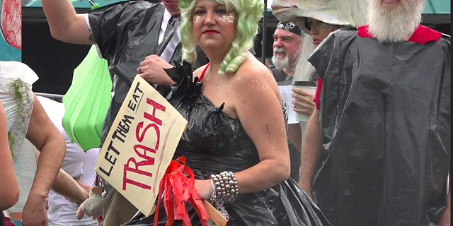 A woman holds a "Let them eat trash" sign while wearing a garbage bag for a dress at an organized 'trash parade' protest.