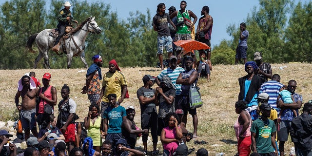 United States Border Patrol agents on horseback watch as Haitian migrants sit on the riverbank near a camp on the banks of the Rio Grande near the Acuna del Rio International Bridge in Del Rio, Texas, on September 19, 2021.