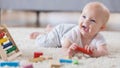 A cute baby lays on her tummy on a rug in a living room and sings with an open mouthed smile.  She is holding one of several wooden blocks.  There is an abacus in the foreground.