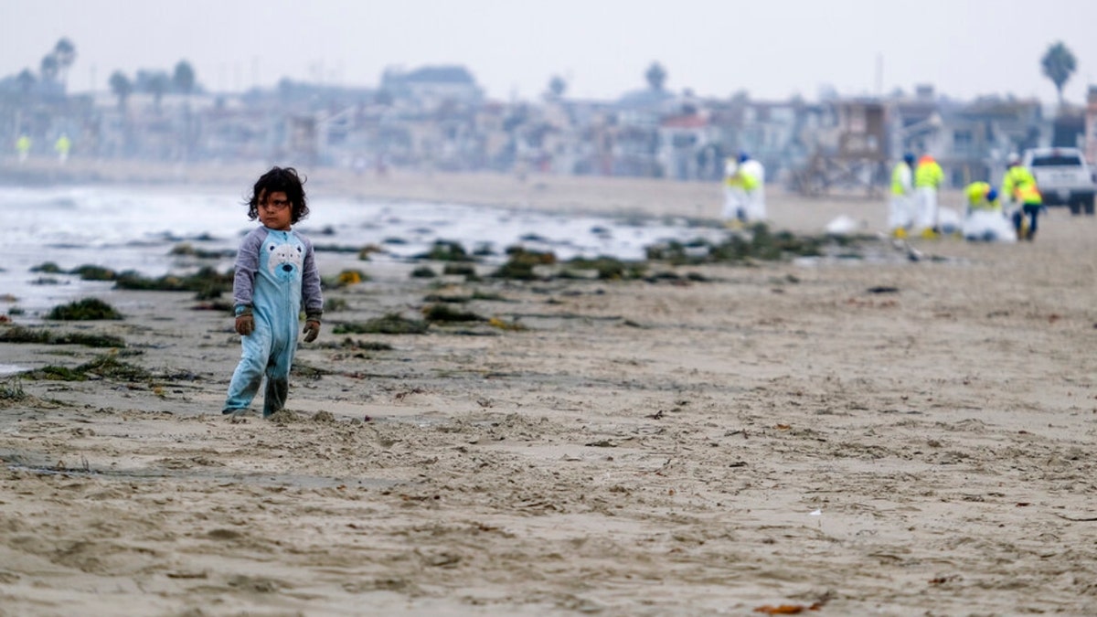 A boy stands on the sands as workers in protective suits clean the contaminated beach after an oil spill in Newport Beach, Calif., on Thursday, Oct. 7, 2021. A major oil spill off the coast of Southern California fouled popular beaches and killed wildlife while crews scrambled Sunday, to contain the crude before it spread further into protected wetlands. 