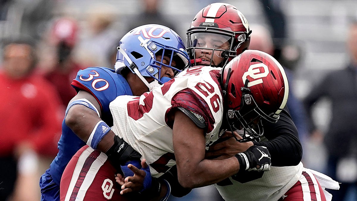 Oklahoma running back Kennedy Brooks (26) hands the ball to quarterback Caleb Williams, back, as he is being tackled by Kansas linebacker Rich Miller (30) during the second half of a game Saturday, Oct. 23, 2021, in Lawrence, Kan. 