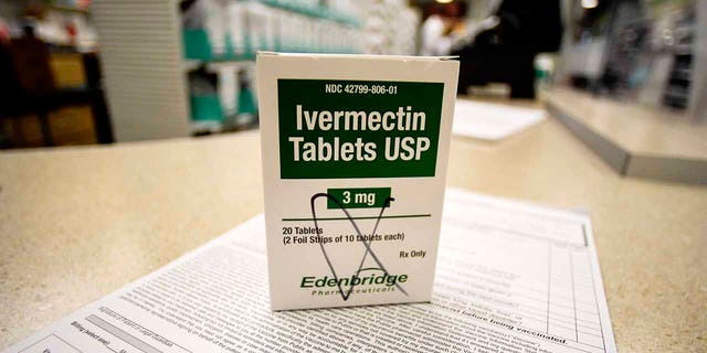 A box of ivermectin is shown in a pharmacy as pharmacists work in the background.