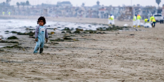 A boy stands on the sands as workers in protective suits clean the contaminated beach after an oil spill in Newport Beach, Calif., on Thursday, Oct. 7, 2021. A major oil spill off the coast of Southern California fouled popular beaches and killed wildlife while crews scrambled Sunday, to contain the crude before it spread further into protected wetlands. 