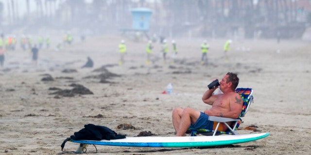A man rests as workers in protective gear continue to clean the contaminated beach in Huntington Beach, Calif. On Monday, October 11, 2021. Huntington Beach reopened its shoreline this morning after water test results returned with undetectable amounts of oil associated with toxins in ocean water, officials from city and state parks in California said.