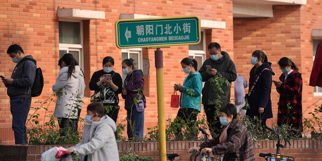 Women wearing face masks to help curb the spread of the coronavirus ride scooters passing by masked residents lined up to receive booster shots against COVID-19 at a vaccination site in Beijing on Monday.