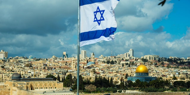 An Israeli flag flying over Jerusalem with the Temple Mount and Al-Aqsa Mosque in the distance.