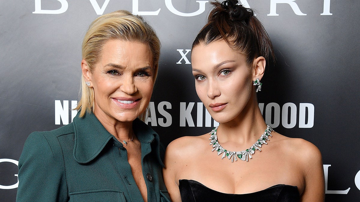 Yolanda Hadid in a dark teal blouse smiles next to daughter Bella Hadid in an up-do and black dress with a diamond necklace