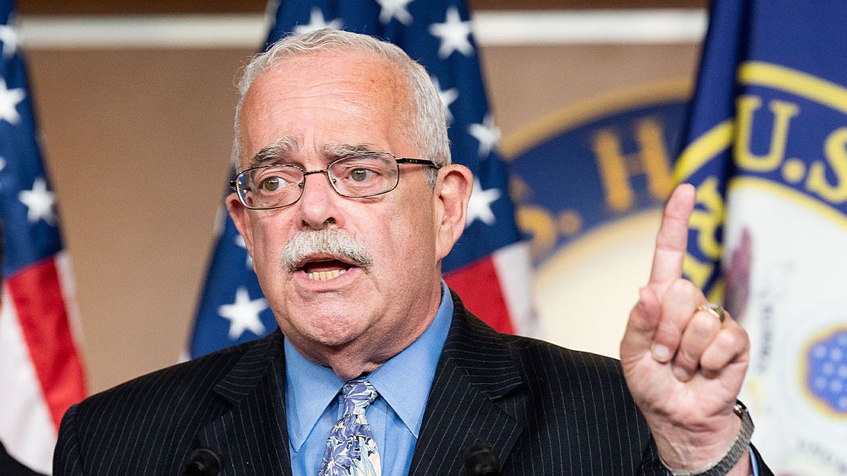 Rep. Gerry Connolly wearing a dark suit, light blue button down while standing in front of 3 flags