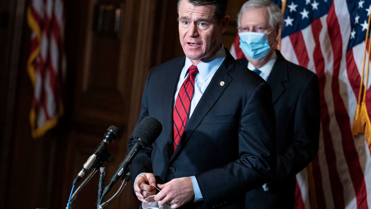 Sen. Todd Young in a suit and red tie speaking in front of Mitch McConnell