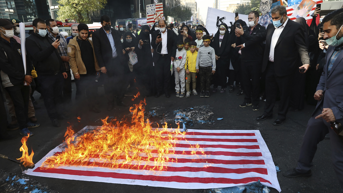 American flag burns on the ground with onlookers surrounding it 