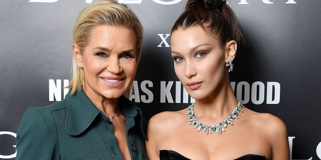 Yolanda Hadid in a dark teal blouse smiles next to daughter Bella Hadid in an up-do and black dress with a diamond necklace