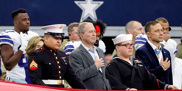 El ex presidente George W. Bush.  Bush se encuentra entre los miembros de las Fuerzas Armadas de los Estados Unidos mientras suena el himno nacional antes de la primera mitad de un partido de fútbol americano de la NFL.