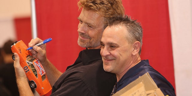 John Schneider autographs a miniature "General Lee," and poses with a fan at the Motor City Comic Con at Suburban Collection Showplace on May 16, 2015 in Novi, Michigan. 