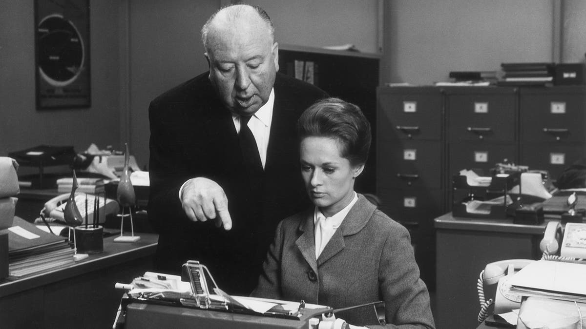 British director Alfred Hitchcock directs American actress Tippi Hedren while she sits in front of a typewriter on the set of his film 