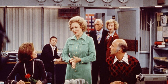 Mary Tyler Moore (as Mary Richards) sits at her desk, links, as Betty White (as Sue Ann Nivens) ,sentrum, talks to her and Gavin MacLeod (as Murray Slaughter) reg sit, in a scene from "Die Mary Tyler Moore Show" in the mid-1970s. Behind them, to the right, are Ted Knight (1923 - 1986) (as Ted Baxter) and Georgia Engel (as Georgette Franklin Baxter).