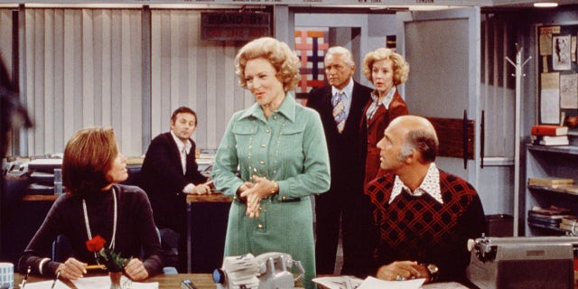 Mary Tyler Moore (als Mary Richards) zit aan haar bureau, Betty White (als Sue op Nivens) in het midden, pratend met haar en Gavin McLeod (als Murray Slaughter), die aan de rechterkant zitten. "Mary Tyler Moore Show" Midden jaren 70. Rechts achter hen staan Ted Knight (1923 - 1986) (als Dead Baxter) en Georgia Angel (als Georgette Franklin Baxter).