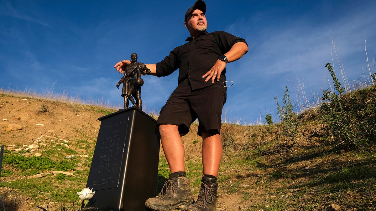 Artis Dan Medina poses next to his work, a bronze sculpture honoring former Los Angeles Lakers NBA basketball player Kobe Bryant, his daughter Gianna Bryant, and the names of those who died, at the site of a 2020 helicopter crash in Calabasas, Calif, on Wednesday, Jan. 26, 2022. 