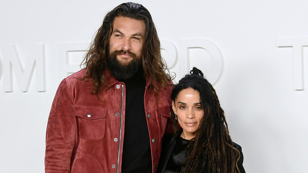Jason Momoa in a black turtleneck and red rust jacket poses with Lisa Bonet in a black outfit