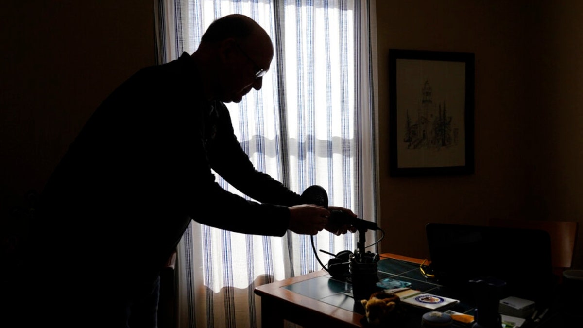 Roger Strukhoff 67, stands in silhouette cleaning off his home office desk Thursday, Jan. 20, 2022, in his DeKalb, Ill., home. Strukhoff was being treated for intestinal bleeding at a hospital outside Chicago this month when he suffered a mild heart attack. Normally, the medical staff would have sent Strukhoff to the intensive care unit, but, overrun with COVID-19 patients, the staff instead had to wheel a heart monitor into his room and quickly administer nitroglycerin and morphine.