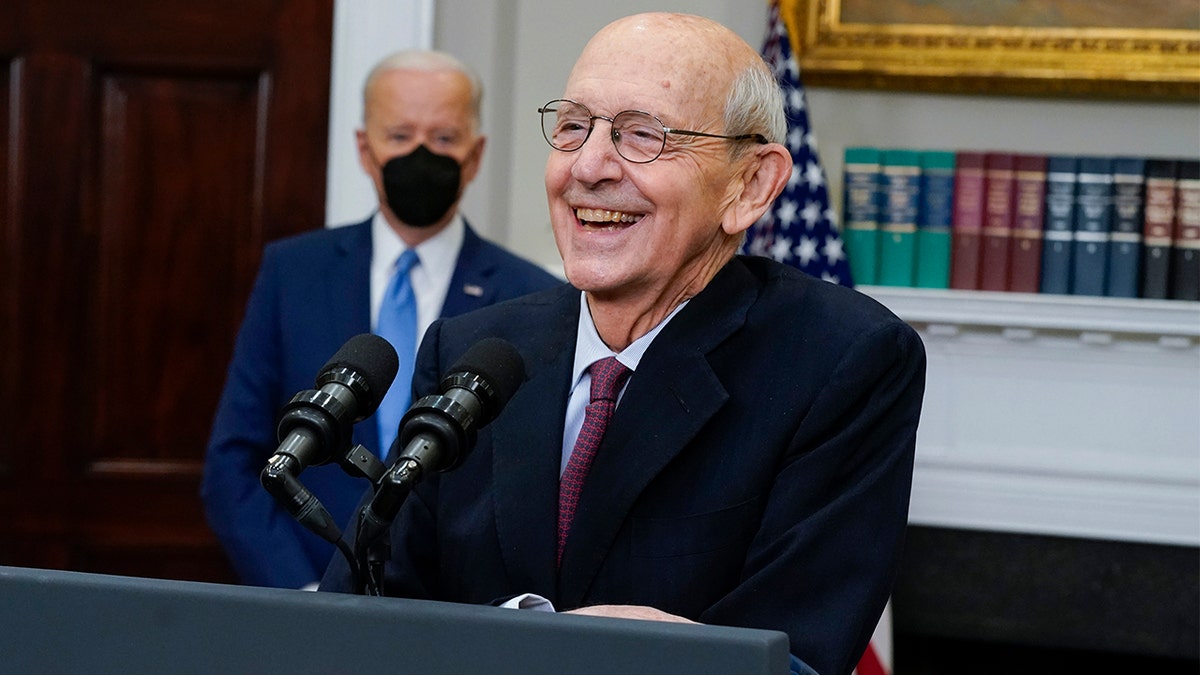 Supreme Court Associate Justice Stephen Breyer at podium laughs in a suit as Biden stands behind him