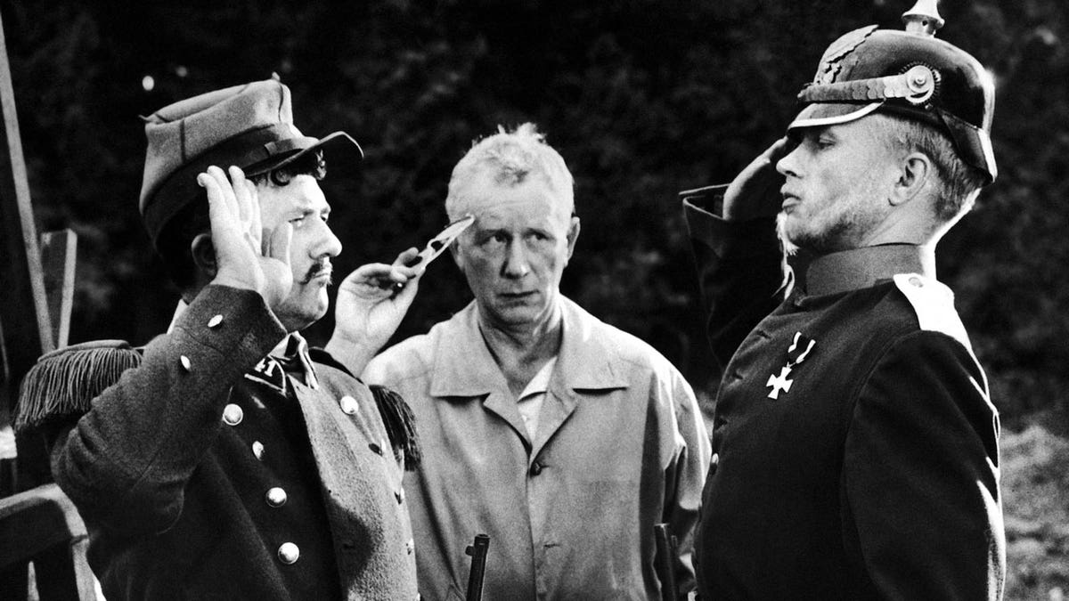 German director Helmut Kaeutner, at center, shows German actor Hardy Krueger, right, and French actor Jean Richard how to salute during filming in France of "Die Gans von Sedan" (Without Trumpet or Drum), a comedy set during the Franco-Prussian War of 1870. 