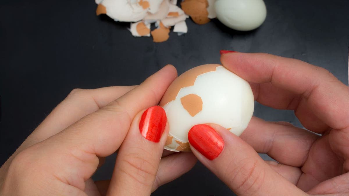 Woman peels a boiled chicken egg with her hands above a dark kitchen counter.