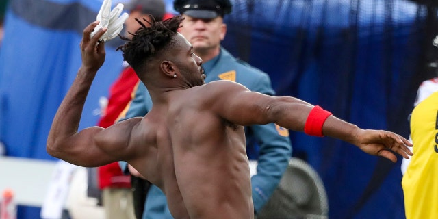 Um soldado da Polícia do Estado de Nova Jersey, ao fundo, observa o wide receiver do Tampa Bay Buccaneers, Antonio Brown (81) joga suas luvas nas arquibancadas enquanto a ofensiva de seu time está em campo contra o New York Jets durante o terceiro quarto de um NFL jogo de futebol no domingo, 2 de janeiro de 2022, em East Rutherford, NJ