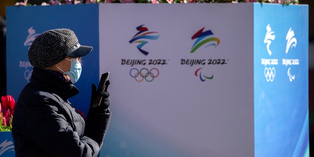 A woman wearing a face mask to protect against COVID-19 sits near landscaping decorated with the logos for the Beijing Winter Olympics and Paralympics on a pedestrian shopping street in Beijing, Saturday, Jan. 15, 2022.
