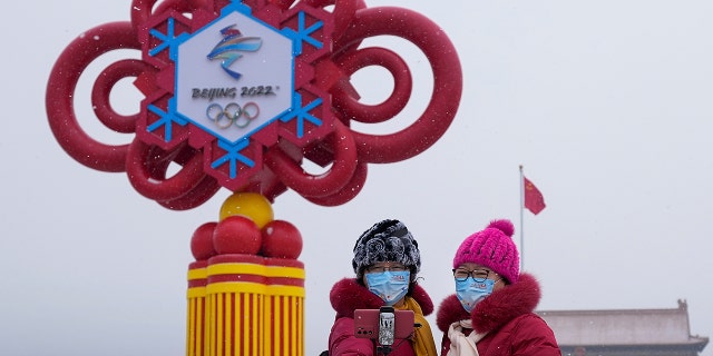 Women wearing face masks printed with a slogan for Beijing Winter Olympics Games take a selfie with a decoration for the Winter Olympics Games on Tiananmen Square in Beijing, 목요일, 1 월. 20, 2022.
