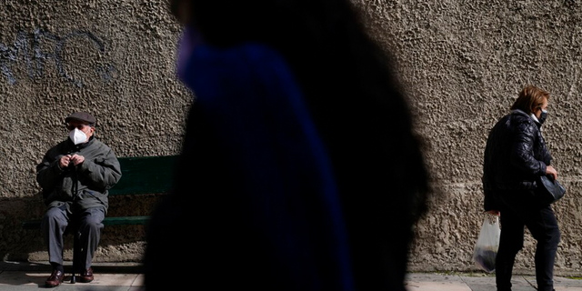 A man wearing a face mask to curb the spread of coronavirus sits on a bench as pedestrians walk outside Evangelismos Hospital in Athens, Griekeland, Maandag, Jan.. 17, 2022.