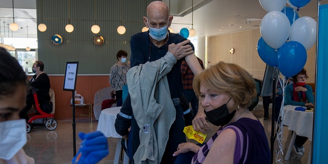 A man pulls his shirt up while his wife receives her fourth dose of the coronavirus vaccine in a private nursing home in Petah Tikva, Israel,Tuesday, Jan. 4, 2022.