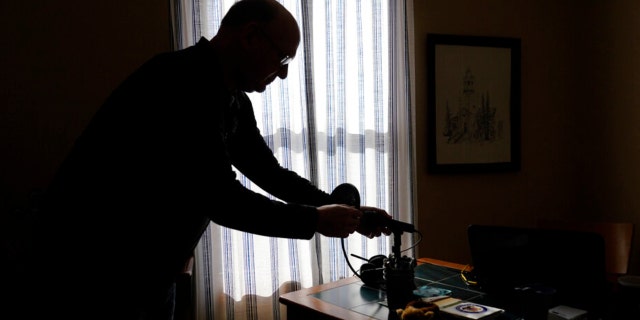 Roger Strukhoff 67, stands in silhouette cleaning off his home office desk Thursday, Jan. 20, 2022, in his DeKalb, Ill., home. Strukhoff was being treated for intestinal bleeding at a hospital outside Chicago this month when he suffered a mild heart attack. Normally, the medical staff would have sent Strukhoff to the intensive care unit, but, overrun with COVID-19 patients, the staff instead had to wheel a heart monitor into his room and quickly administer nitroglycerin and morphine.