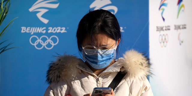 A woman wearing a face mask to protect against COVID-19 sits near landscaping decorated with the logos for the Beijing Winter Olympics and Paralympics on a pedestrian shopping street in Beijing, Sabato, Jan. 15, 2022. 