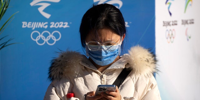 A woman wearing a face mask to protect against COVID-19 sits near landscaping decorated with the logos for the Beijing Winter Olympics and Paralympics on a pedestrian shopping street in Beijing, Saterdag, Jan.. 15, 2022.