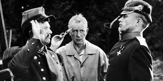 German director Helmut Kaeutner, at center, shows German actor Hardy Krueger, right, and French actor Jean Richard how to salute during filming in France of "Die Gans von Sedan" (Without Trumpet or Drum), a comedy set during the Franco-Prussian War of 1870.