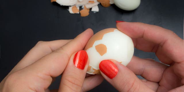 Woman peels a boiled chicken egg with her hands above a dark kitchen counter.