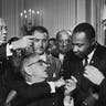 President Lyndon B. Johnson shakes the hand of Dr. Martin Luther King Jr. at the signing of the Civil Rights Act while officials look on in Washington D.C. 