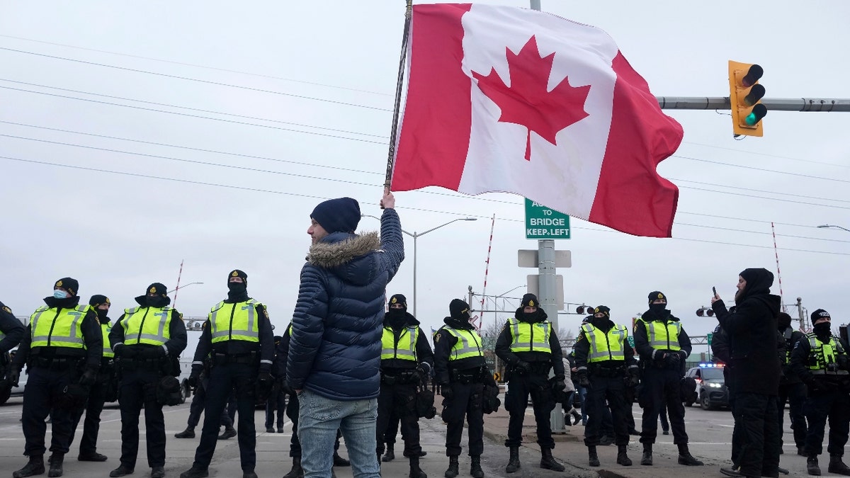 A supporter waves a Canadian flag on a hockey stick as police officers enforce an injunction against their demonstration, which has blocked traffic across the Ambassador Bridge by protesters against COVID-19 restrictions, in Windsor, Ont., Saturday, Feb. 12, 2022. THE CANADIAN PRESS/Nathan Denette