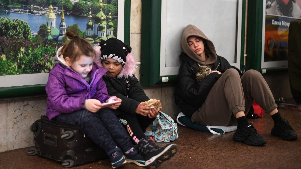Lia (L) and Jasmine (C) use a phone as they take shelter with their parents (unseen) in a station in central Kyiv on February 24, 2022.