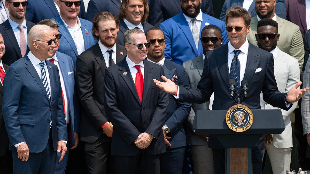 Tampa Bay Buccaneers quarterback Tom Brady speaks alongside President Biden and team owner Bryan Glazer (center) during a ceremony honoring the Tampa Bay Buccaneers for their Super Bowl LV Championship on the South Lawn of the White House in Washington, D.C., on July 20, 2021.