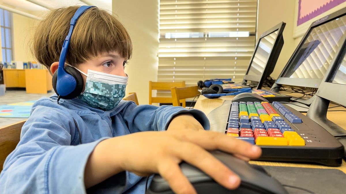 Caucasian Little boy using a computer at his school library wearing a protective face mask