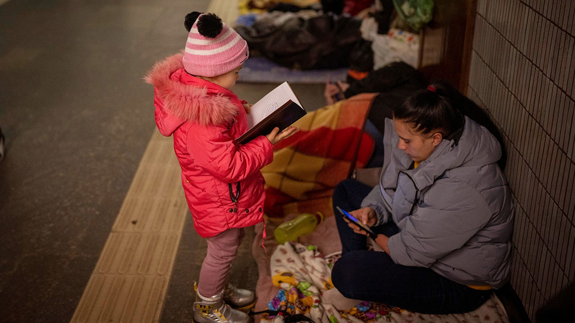 A girl looks at a notebook next to her mother as they stand in the Kyiv subway, using it as a bomb shelter, in Ukraine, Saturday Feb. 26, 2022.