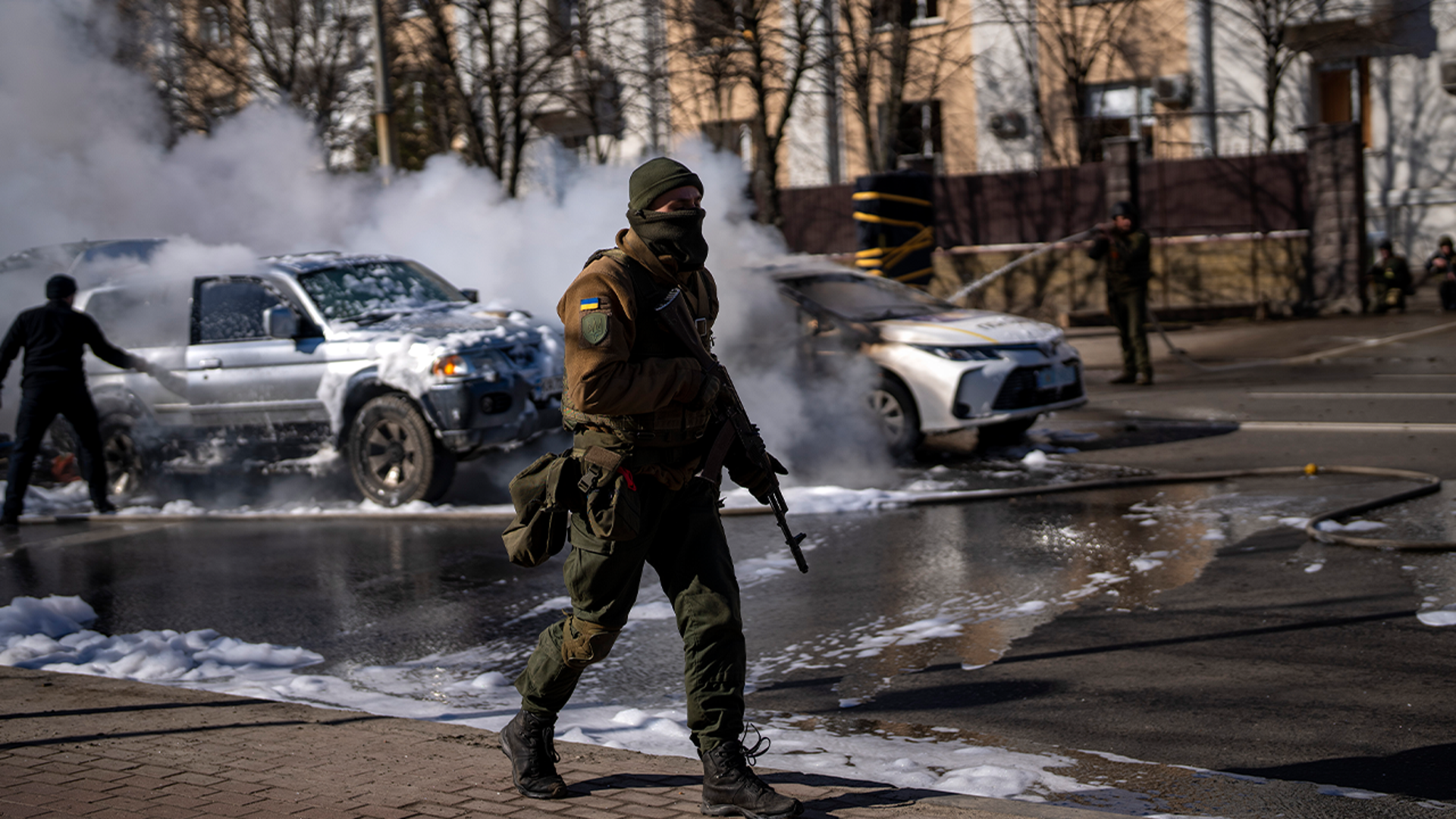Ukrainian soldiers take positions outside a military facility as two cars burn, in a street in Kyiv, Ukraine, Saturday, Feb. 26, 2022. Russian troops stormed toward Ukraine's capital Saturday, and street fighting broke out as city officials urged residents to take shelter.