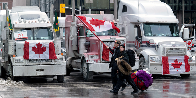 Protesters walk with bags past trucks parked on downtown streets on Wednesday, Feb. 2, 2022, in Ottawa, Ontario. Thousands of protesters railing against vaccine mandates and other COVID-19 restrictions descended on the capital, deliberately blocking traffic around Parliament Hill.