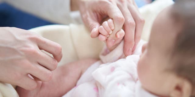 A baby holds a dad's finger in this image. Wrote a new mom on social media as she asked for others' opinions, "He shouldn't be gone even a day away from our baby" (not pictured). 