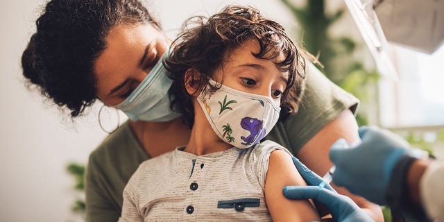 A young child is seen wearing a mask while getting a vaccine.