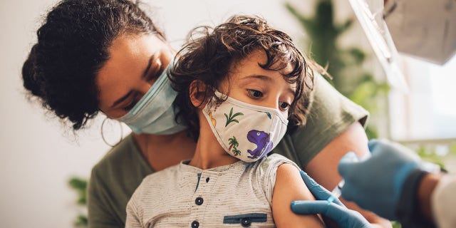 Cute boy wearing face mask taking vaccine at home. Kid with mother receiving a COVID-19 vaccine from a healthcare worker at home.