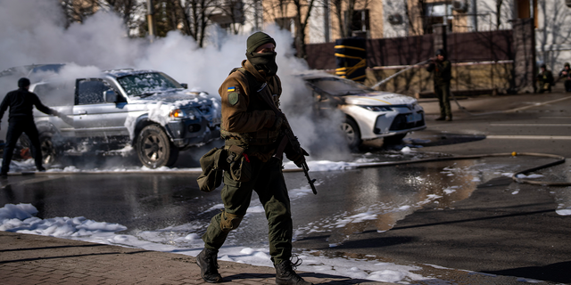 Soldados ucranianos toman posición frente a una instalación militar mientras dos autos se queman, en una calle de Kiev, Ucrania, el sábado 26 de febrero de 2022. Las tropas rusas irrumpieron en la capital ucraniana el sábado y estallaron las protestas callejeras cuando los funcionarios de la ciudad instaron a los residentes a refugiarse.  . 